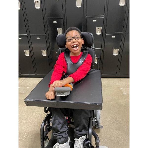 young child in wheelchair with arm on device in front to control the chair; kid is wearing glasses and a red shirt