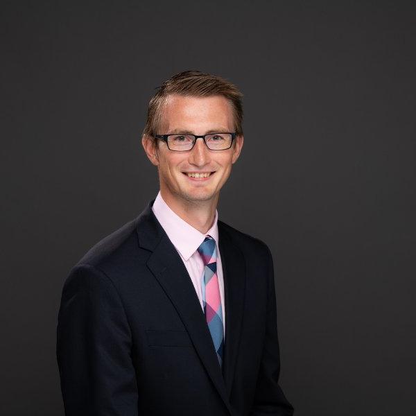 Zachary DeBruine seated for a headshot in a dark suit, colorful tie